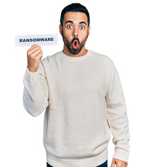 Young hispanic man with beard holding ransomware banner scared and amazed with open mouth for surprise, disbelief face