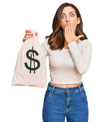 Young brunette woman holding dollars bag covering mouth with hand, shocked and afraid for mistake....