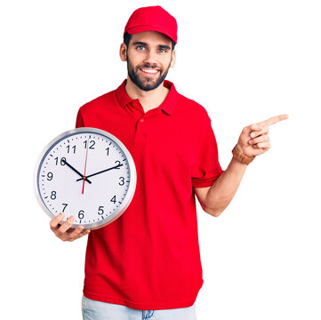 Young handsome man with beard wearing delivery uniform holding big clock smiling happy pointing with hand and finger to the side