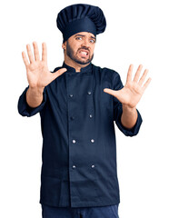 Young hispanic man wearing cooker uniform afraid and terrified with fear expression stop gesture with hands, shouting in shock. panic concept.