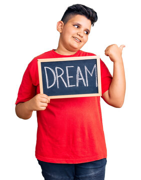 Little boy kid holding blackboard with dream word pointing thumb up to the side smiling happy with open mouth