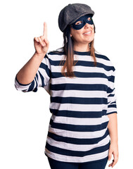 Young beautiful brunette woman wearing burglar mask pointing finger up with successful idea. exited and happy. number one.