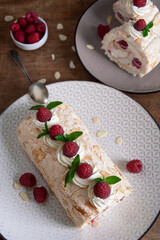 Top view of a meringue roll with raspberries and almond petals, decorated with cream, raspberries and mint leaves, on a wooden background. Vertical