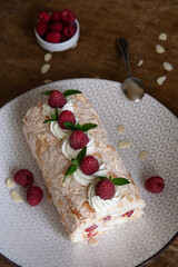Top view of a meringue roll with raspberries and almond petals, decorated with cream, raspberries and mint leaves, on a wooden background. Vertical