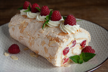 Side view of a meringue roll with raspberries and almond petals, decorated with cream, raspberries and mint leaves, on a dark wooden background