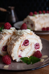 Two pieces of meringue roll with cream, raspberries and almond petals. A whole roll and a coffee pot in the background. Black background. Close-up. Vertical