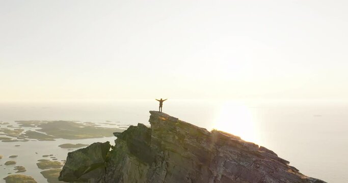 Happy climber lift arms celebrate on Norway mountain top in midnight sun; drone
