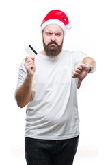 Young caucasian man wearing christmas hat holding credit card over isolated background with angry face, negative sign showing dislike with thumbs down, rejection concept