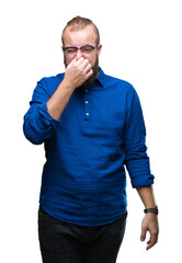 Young caucasian hipster man wearing glasses over isolated background smelling something stinky and disgusting, intolerable smell, holding breath with fingers on nose. Bad smells concept.