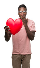 Young african american man holding red heart cover mouth with hand shocked with shame for mistake, expression of fear, scared in silence, secret concept