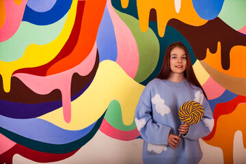 Portrait of teen lady model with round giant lollipop at colored wall, smiling looking at camera. Teenage cover girl in blue fluffy sweater with candy on stick. Sweet life concept. Copy ad text space