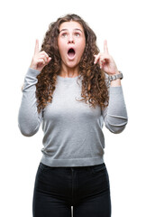 Beautiful brunette curly hair young girl wearing a sweater over isolated background amazed and surprised looking up and pointing with fingers and raised arms.