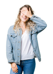 Beautiful young blonde woman wearing denim jacket over isolated background smiling and laughing with hand on face covering eyes for surprise. Blind concept.