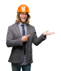 Young handsome architec man with long hair wearing safety helmet over isolated background amazed and smiling to the camera while presenting with hand and pointing with finger.