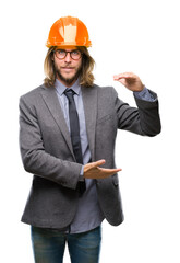 Young handsome architec man with long hair wearing safety helmet over isolated background gesturing with hands showing big and large size sign, measure symbol. Smiling looking at the camera