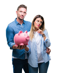 Young couple in love holding piggy bank over isolated background serious face thinking about...