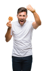 Young handsome man eating a sweet waffle over isolated background annoyed and frustrated shouting with anger, crazy and yelling with raised hand, anger concept