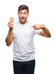 Young handsome man eating chocolate chips cookie over isolated background with surprise face pointing finger to himself