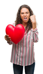 Fototapeta na wymiar Young hispanic woman in love holding red heart annoyed and frustrated shouting with anger, crazy and yelling with raised hand, anger concept