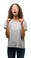 Beautiful young hispanic woman wearing glasses amazed and surprised looking up and pointing with fingers and raised arms.