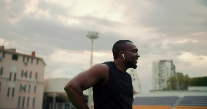 A Black-skinned man in a black sports uniform walks through the city stadium and rests looking back against the gray sky