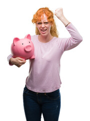 Young redhead woman holding piggy bank annoyed and frustrated shouting with anger, crazy and yelling with raised hand, anger concept