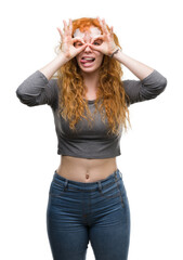 Young redhead woman doing ok gesture like binoculars sticking tongue out, eyes looking through fingers. Crazy expression.