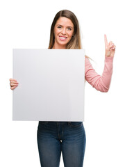 Beautiful young woman holding advertising banner surprised with an idea or question pointing finger with happy face, number one