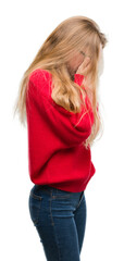 Blonde teenager woman wearing red sweater with sad expression covering face with hands while crying. Depression concept.