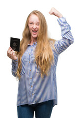 Blonde teenager woman holding passport of Unites States of America annoyed and frustrated shouting with anger, crazy and yelling with raised hand, anger concept