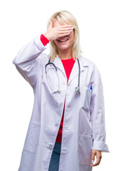 Young beautiful blonde doctor woman wearing medical uniform over isolated background smiling and laughing with hand on face covering eyes for surprise. Blind concept.