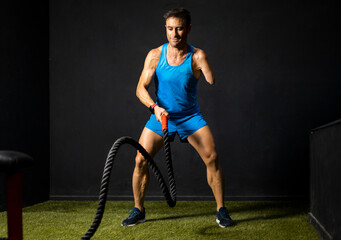 Horizontal photo of an adult athlete with an amputated arm performing a battle rope exercise inside...