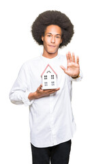 Young african american man with afro hair holding house with open hand doing stop sign with serious and confident expression, defense gesture