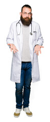 Young blond doctor man with beard wearing medical coat clueless and confused expression with arms and hands raised. Doubt concept.