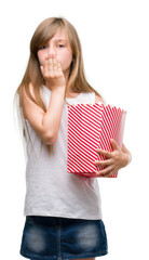 Young blonde toddler holding popcorn pack cover mouth with hand shocked with shame for mistake, expression of fear, scared in silence, secret concept