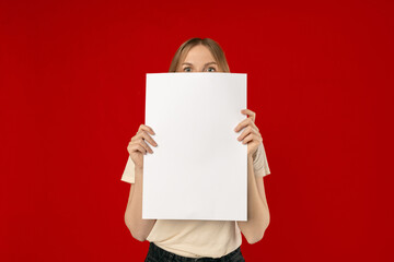 Smiling Caucasian woman holding empty vertical white poster advertising something standing on red studio background. Mockup