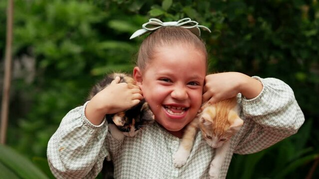 Ni&ntilde;a linda latina rubia feliz con una gran sonrisa acariciando jugando con dos mascotas gatitos gatos felinos traviesos e inquietos y curiosos en el jard&iacute;n al exterior al aire libre un d&iacute;a lluvioso 