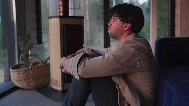 Depressed young man sitting on the floor by the sofa in the living room against the background of the fireplace. 