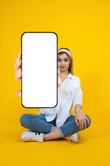 Showing mobile phone mock up, full body view sitting blonde girl showing mobile phone mock up. Isolated yellow background. White blank empty screen smartphone workspace. App, web page recommending.
