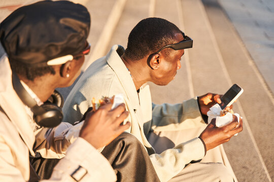 Multiracial hipster guys eating fast food and using smartphone while sitting on street stairs