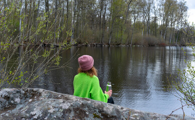 Young female in green sweater and hat sit on rock resting, looking at the lake while drinking water from metal bottle. Mindfulness in nature. Cold weather lake shore.