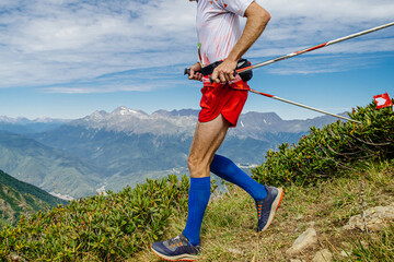 male athlete running from mountainside in compression socks on his feet and trail trekking poles in...