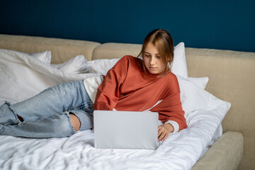 Thoughtful teen girl lies relaxed on bed with laptop typing on keyboard communicates on social networks chooses college for admission. Internet addiction among teenagers, students lack of interests
