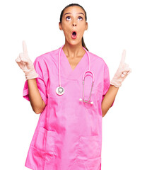 Young hispanic woman wearing doctor uniform and stethoscope amazed and surprised looking up and pointing with fingers and raised arms.