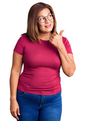 Middle age latin woman wearing casual clothes and glasses smiling with happy face looking and pointing to the side with thumb up.