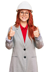 Young redhead woman wearing architect hardhat success sign doing positive gesture with hand, thumbs up smiling and happy. cheerful expression and winner gesture.