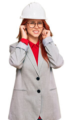 Young redhead woman wearing architect hardhat covering ears with fingers with annoyed expression for the noise of loud music. deaf concept.