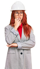 Young redhead woman wearing architect hardhat looking stressed and nervous with hands on mouth biting nails. anxiety problem.