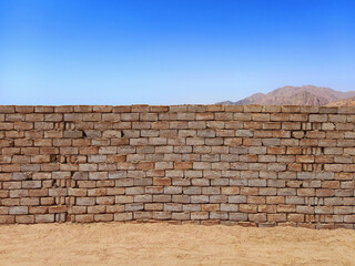 Brick wall in the sand of the Dahab desert. Stone fence under blue sky and arid mountains of Egypt. Vintage arabic mud wall in the desert made with beach rocks and coral stones