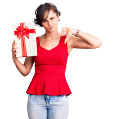 Fototapeta premium Beautiful young woman with short hair holding gift with angry face, negative sign showing dislike with thumbs down, rejection concept